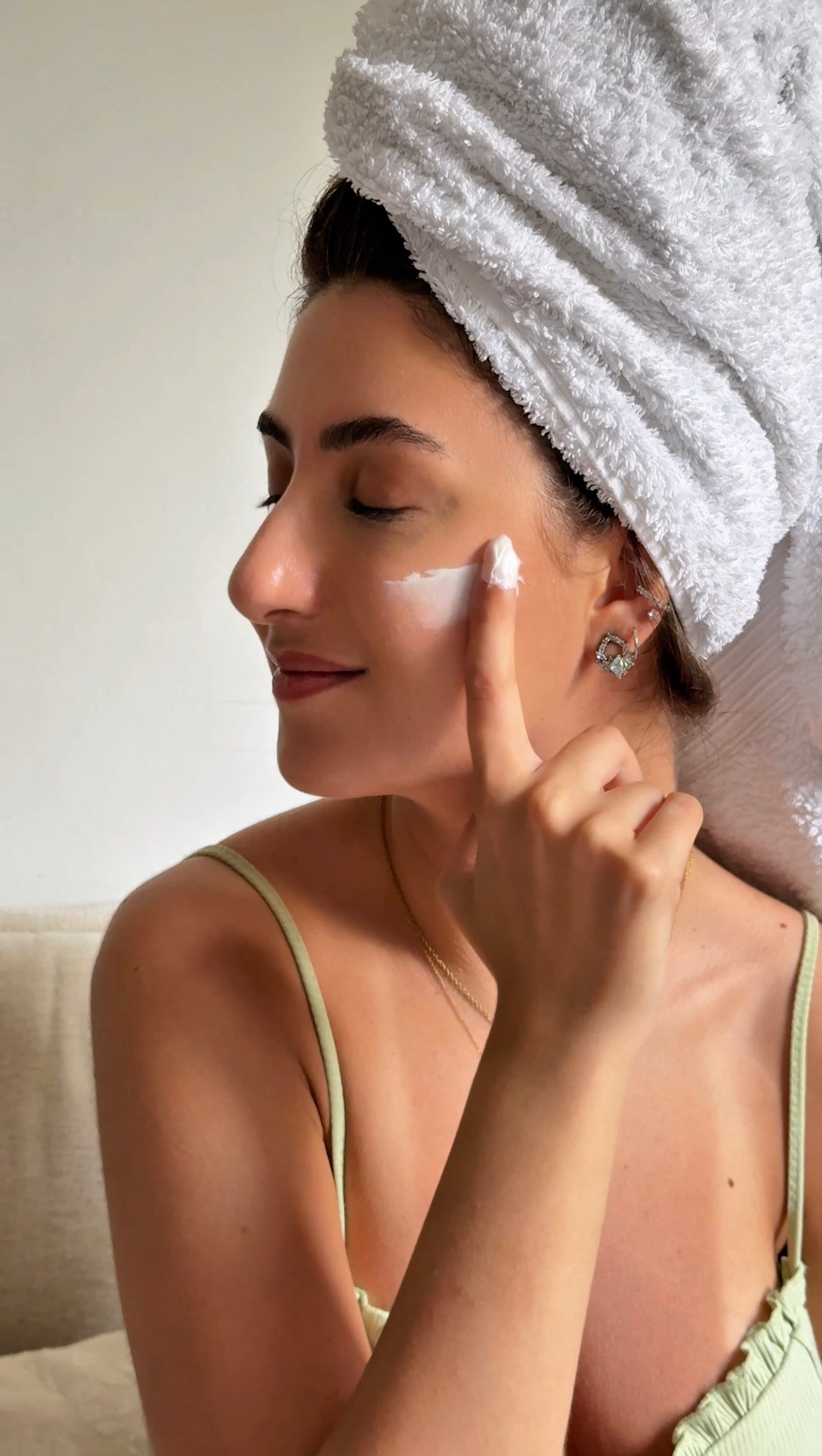 Woman applying cream to her face with a towel on her head, indoors.