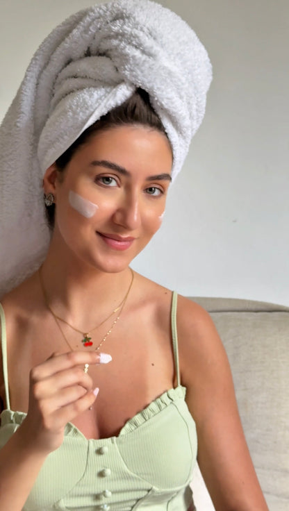 Woman with a towel wrapped around her head, applying cream to her face.
