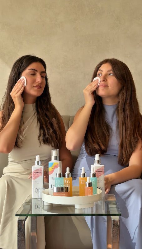 Two women applying a facial product with skincare products on a table in front of them.